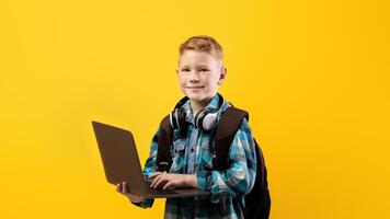 Technologies in school education. Cheerful schoolboy with backpack and headset holding laptop over yellow studio background, copy space. Cute kid using laptop for studying, doing homework photo