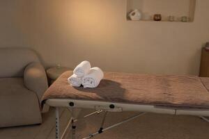 Minimalist spa interior with a clean massage table topped with neatly rolled white towels in a calming environment ready for a wellness treatment session. photo