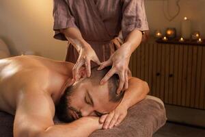 Professional therapist performing a relaxing head massage on a bearded man lying on a spa table in a calm, candle-lit wellness room with warm and peaceful atmosphere. photo