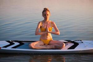 Woman in yellow bikini practicing yoga on a paddleboard at sunrise, meditating in lotus pose over calm water, symbolizing balance, peace, and healthy lifestyle. photo