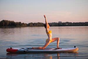 Woman in yellow bikini practicing yoga on paddleboard in warrior pose at sunrise, balancing on calm water, expressing focus, strength, and harmony with nature. photo