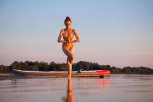 Woman in yellow bikini practicing yoga in tree pose on shallow water beside paddleboard at sunrise, expressing balance, mindfulness, and connection to nature. photo