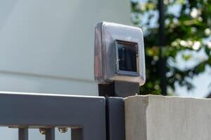 Solar-powered gate light is positioned atop a gray metal fence, providing illumination during nighttime hours. The device absorbs sunlight during the day for energy photo