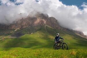 View of cyclist standing with mountain bike in summer green fields in Dolomites with high rocky mountain in background in thick clouds on sunny day. Off-piste riding photo