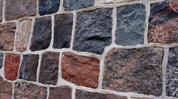 Close-up view of a multicolored stone wall with irregular rectangular blocks and white grout. The textured surface and natural colors give a rustic and architectural feel. photo