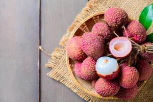 fresh ripe lychees in a bamboo basket with green leaves on a rustic wooden table. One peeled lychee reveals the juicy white flesh inside photo