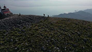 Hikers climb up the rocky mountainside approaching a small church on its top enjoying the picturesque panoramic view and joyfully throwing their hands up, aerial view video