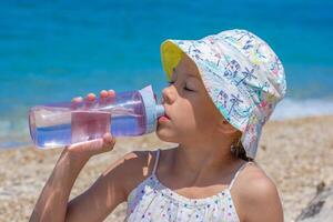Caucasian child of 8 9 years in a vibrant sun hat is sipping water from a transparent bottle on a sunny beach with closed eyes photo