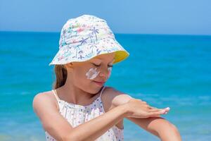Child is applying sunscreen on her arm at the beach, wearing a colorful hat, with bright ocean waves in the background, emphasizing sun protection and enjoyment photo