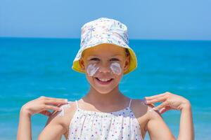 Child happily applying sunscreen on her face and shoulders at the beach, wearing a vibrant hat, with the ocean waves and bright sky creating a joyful summer atmosphere photo