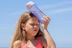 Child of 8 9 years with closed eyes holding bottle of water on forehead feeling headache because of heatstroke in summer over blue sky photo
