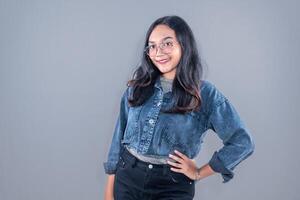 A young woman stands confidently against a gray background, smiling warmly. She is wearing a cropped denim jacket, black high-waisted pants, and round glasses. Her long, wavy hair frames her face photo