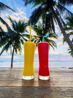 Two vibrant, tall glasses filled with colorful tropical drinks, one bright yellow and one vivid red, stand on a rustic wooden table. In the background, silhouetted palm trees frame a serene beach photo
