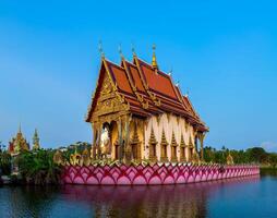 A stunning, highly ornate Thai temple with golden gables and red tiled roofs stands majestically on a vibrant pink lotus petal-shaped base, appearing to float on a calm body of water. The temple's photo