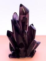 A striking, eye-level close-up captures a raw, deep purple amethyst crystal cluster, showcasing its natural facets and sharp points. The translucent mineral stands prominently against a soft, light photo