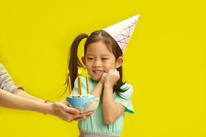 Mood of Holiday. Thoughtful Child Girl in Party Paper Hat Gazes at Birthday Candles on a Cake, Ready to Make Wish and Ready to Blow them out having in Anticipation Staning on Sunny Yellow Background. photo