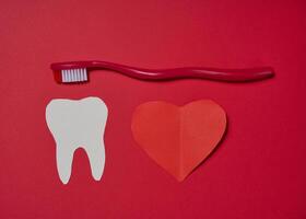 Red toothbrush, white paper tooth cut out and red paper heart placed on red background. photo