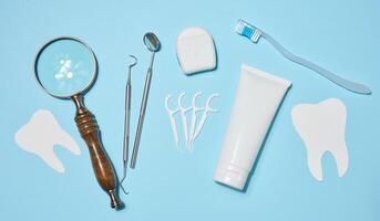 Set of oral hygiene products and tools, tube of toothpaste, dental floss, dental instruments, magnifying glass with heart symbol and paper teeth, blue background. Top view. photo