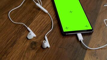 Close-up of Modern Smartphone with Green Screen and White Earbuds on Wooden Surface in Cozy Setting video
