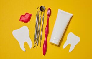 Red toothbrush, white tube of toothpaste, red decorative lips, dental instruments, dental floss in a case and white paper teeth cut out on a yellow background. photo