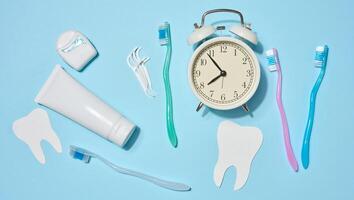 White tube of toothpaste, dental floss in a case, toothbrushes of different colors, white alarm clock, plastic flossers placed on a blue background. Top view. photo