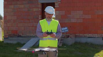 Drone operator controls an aircraft, holding remote controller. Professional builder in helmet and vest standing in front of construction site. House building. Business, real estate and investment video