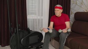 Old grey-haired senior grandfather model doing weight lifting dumbbells exercises in room at home. Retro style Caucasian mature elderly man warming up while sitting on sofa. Healthy sport lifestyle video