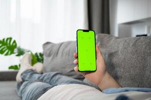 Man using smarphone with green screen lying on the couch at home. Modern technologies,social media,dating and delivery apps concept photo