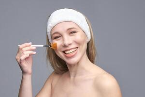 Smiling caucasian female adult applying makeup with brush on cheek with headband on gray background. photo