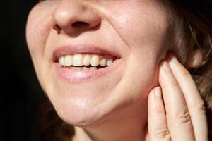 Close-up of young caucasian female smiling and touching cheek with healthy teeth in bright light. photo