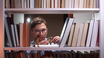 A man in glasses looking at a book shelf with a dinosaur video