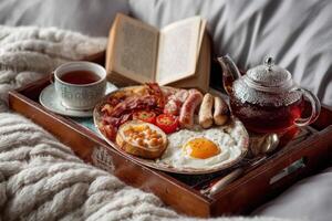 A beautifully arranged breakfast tray includes eggs, bacon, sausages, tomatoes, tea, and a book resting on a cozy blanket photo
