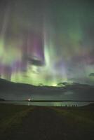Amazing aurora borealis over the countryside of West Iceland with a lighthouse illuminating the surroundings. A palette of colors spread across the Icelandic sky as northern lights photo