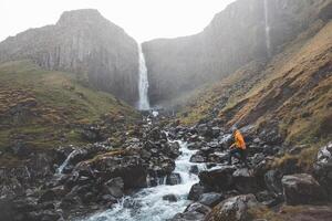 Traveler in an orange jacket walking to Grundarfoss waterfall on the Snaefellsnes Peninsula, Iceland, as mist rises from the cascading water against rugged basalt cliffs photo