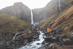 Tourist in an orange jacket stands in awe before Grundarfoss waterfall on the Snaefellsnes Peninsula, Iceland, as mist rises from the cascading water against rugged basalt cliff photo