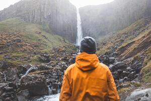 Adventurer in an orange jacket stands in awe before Grundarfoss waterfall on the Snaefellsnes Peninsula, Iceland, as mist rises from the cascading water against rugged basalt cliffs photo