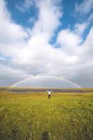 Man stands with arms wide open on a lush green meadow under a rainbow arch in Iceland. The dramatic clouds and blue sky create a magical, uplifting atmosphere. Snaefellsnes peninsula, Iceland photo