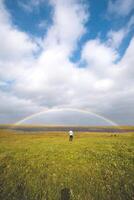 Person stands with arms wide open on a lush green meadow under a rainbow arch in Iceland. The dramatic clouds and blue sky create a magical, uplifting atmosphere. Snaefellsnes peninsula photo