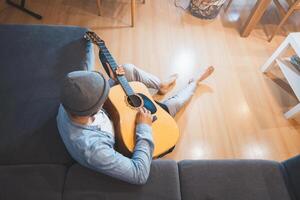 Boy sits on a gray couch, casually strumming an acoustic guitar, immersed in a moment of relaxation and introspection, creating a peaceful and creative atmosphere. Top view photo