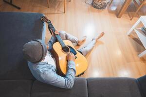 Man sits on a gray couch, casually strumming an acoustic guitar, immersed in a moment of relaxation and introspection, creating a peaceful and creative atmosphere. Top view photo
