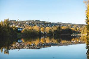Autumn reflections dance on the still waters of Holmendammen Lake in Oslo, surrounded by colorful trees and hillside homes under a crisp, cloudless sky photo
