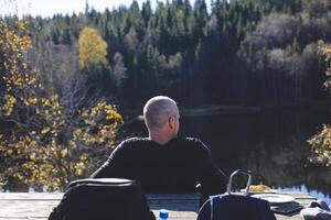 50 years old man relaxes by a forest lake in Oslo Marka, enjoying lunch under golden trees and sunlight, surrounded by still water and deep Scandinavian calm photo