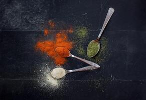 Spoons with spices like paprika, dill, and garlic powder on a dark textured surface top view photo