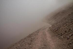A view of a dirt path along a rocky slope covered in a thick layer of dense fog and mist air photo