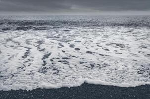 Ocean waves crashing on a dark beach under a cloudy sky creating white foam on the water surface photo