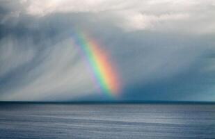 A rainbow over the ocean under a cloudy sky with visible rain and light breaking through the clouds photo