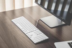 A minimalist workspace featuring a keyboard, monitor, and laptop on a wooden desk surface near a window photo