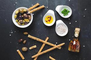 Overhead shot of olives breadsticks and condiments on a dark surface creating a food still life photo