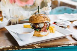 A delicious burger with bacon and cheese served with fries and sauce on a white square plate on a table photo