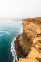 Person standing on a cliff overlooking the ocean with waves crashing against the rocky shore below it photo